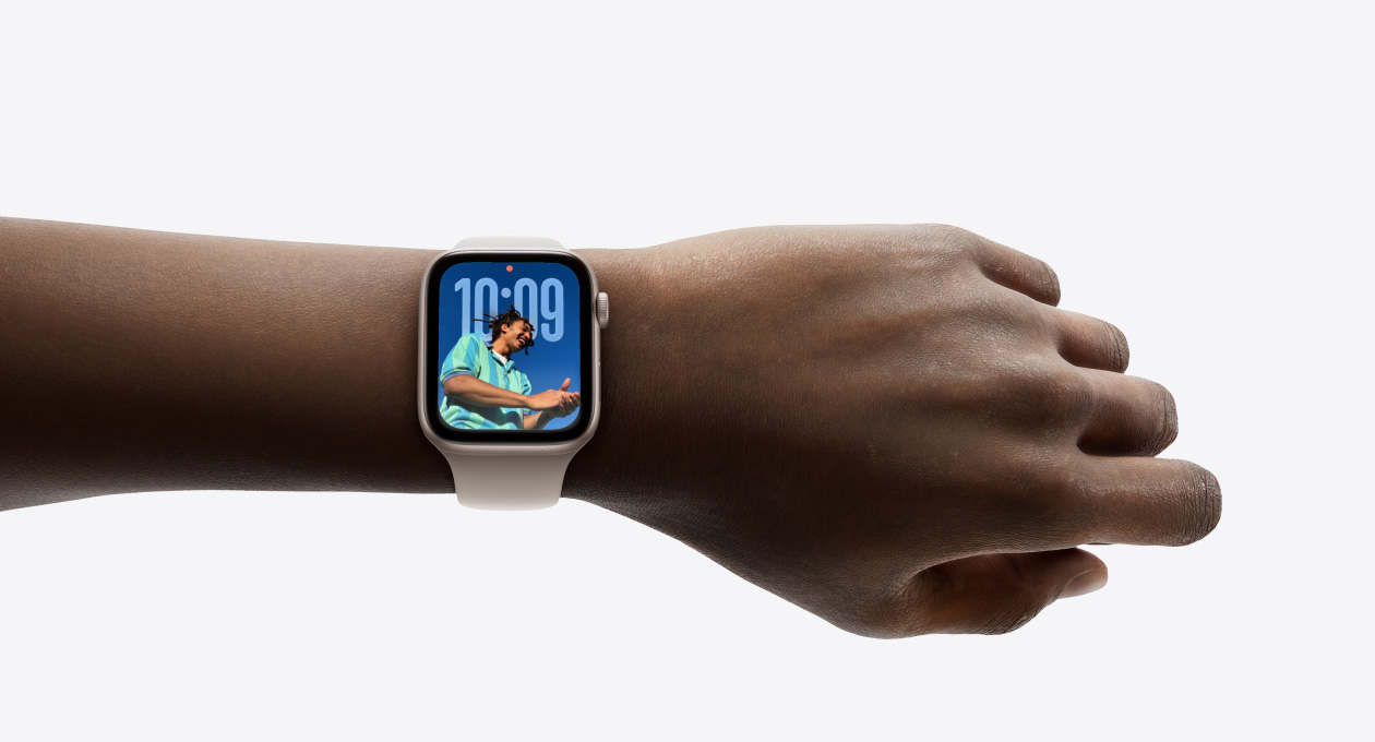 Primer plano de la muñeca de una persona con un Apple Watch en color plata y correa de goma, en cuya pantalla se puede apreciar la app Hora en el nuevo diseño con Liquid Glass