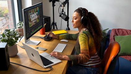 Une femme assise à son bureau chez elle travaille sur son MacBook Pro connecté à un écran externe, elle porte un casque et parle dans un micro