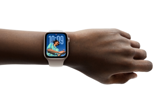 Közeli kép egy ezüstszínű, gumiszíjas Apple Watchról a felhasználó csuklóján. A kijelzőn az Idő app látható az új Liquid Glass-megjelenéssel.