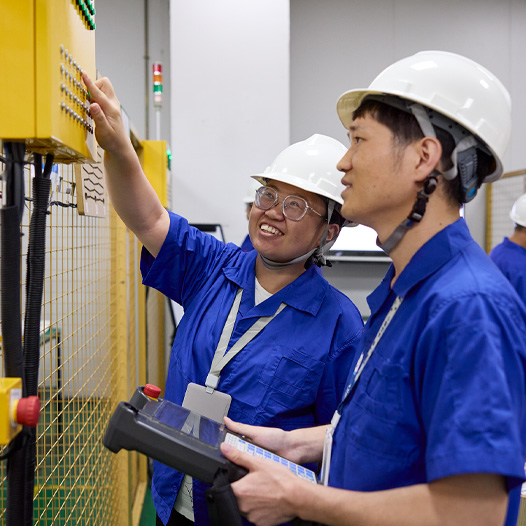 Two employees wearing safety hats and uniform shirts stand by a work cage control box, where one points out a button to the other